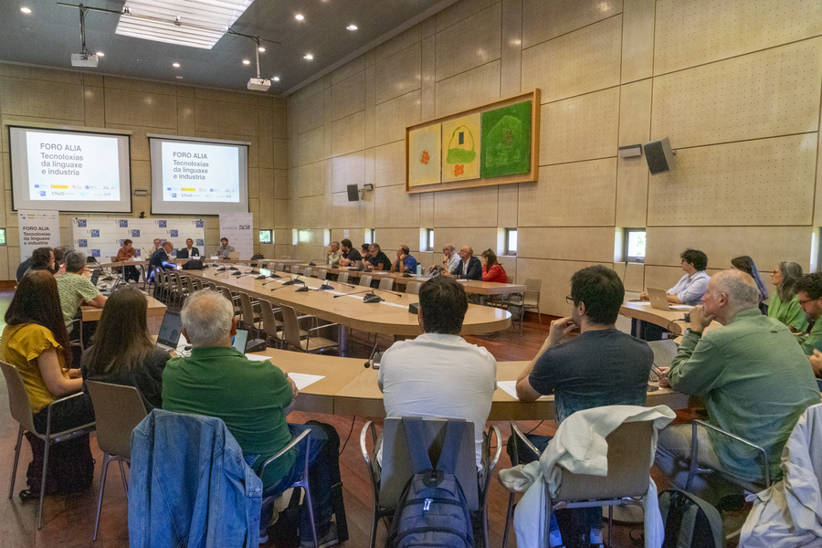 Un grupo de persoas está sentado ao redor dunha mesa con documentos nela, o que suxire que poden estar participando nunha discusión ou reunión. A sala está enchida de luz natural que entra polas grandes ventáns que están detrás deles, creando un ambiente de aberto e colaboración.